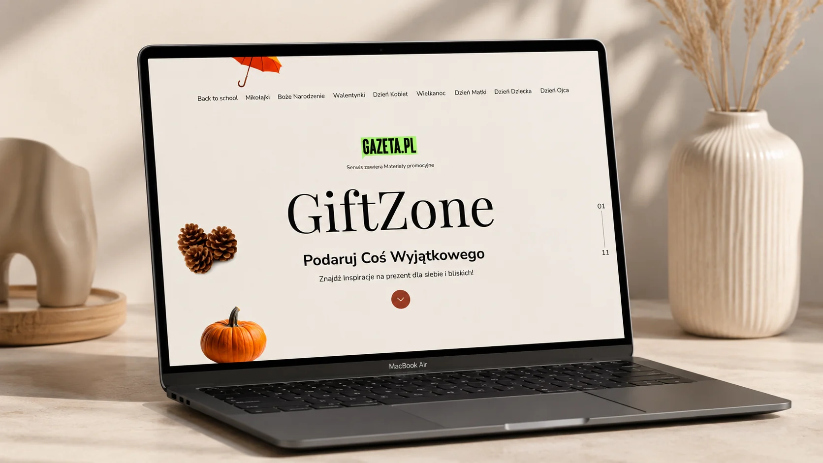 Makieta strony głównej serwisu GiftZone (Gazeta.pl) na laptopie MacBook Air stojącym na jasnym biurku z szyszkami i dynią; nagłówek „GiftZone – Podaruj Coś Wyjątkowego”.